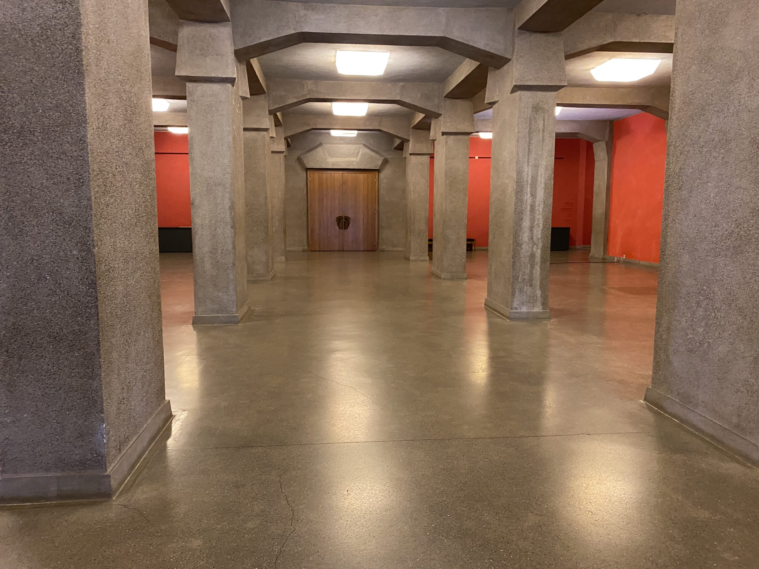 A hall in the Goetheanum with many pillars leading to big doors, red walls and concrete floor & pillars 